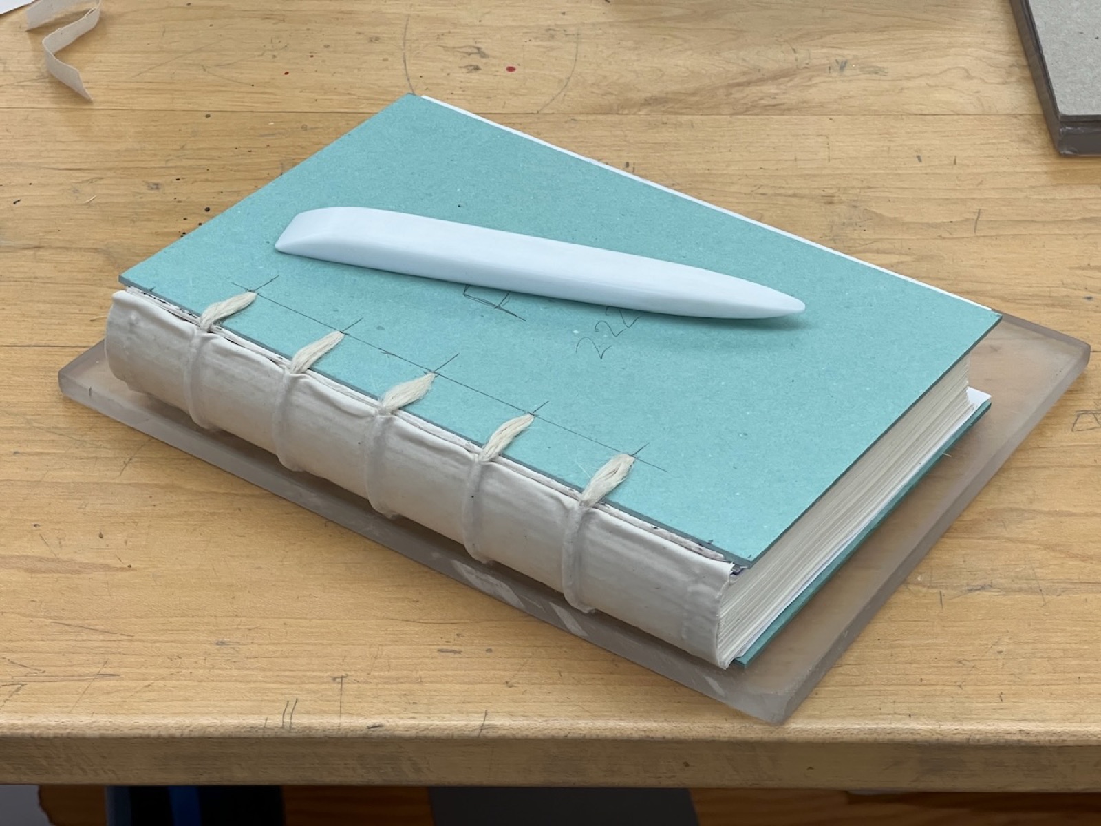 book on table ready for binding, showing laced-in cords into the green cover boards with a teflon bone folder sitting on the book