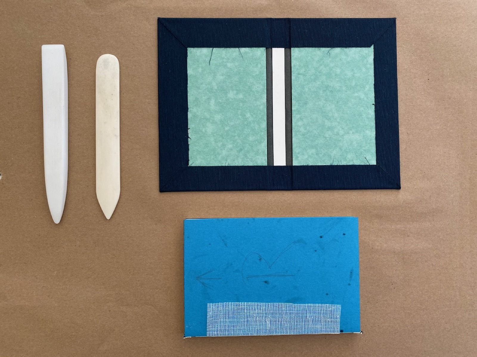 Bone folders beside the flat components of a small case: two covered boards joined by a spine piece.