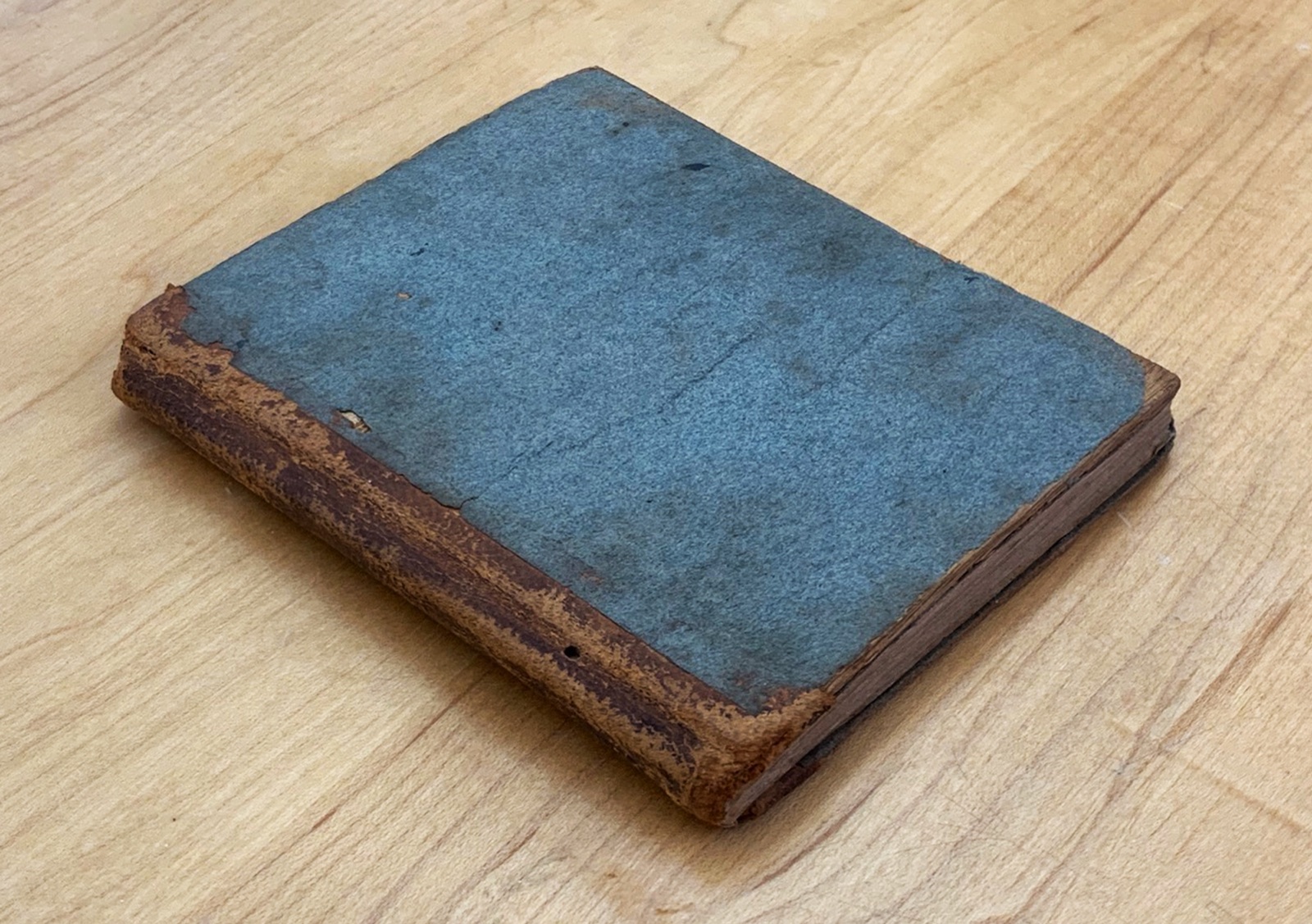 A small antique primer before treatment: worn blue paper boards and exposed, damaged spine.