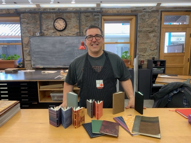 Bob standing behind a table of finished flatback notebooks in a stone-walled classroom.