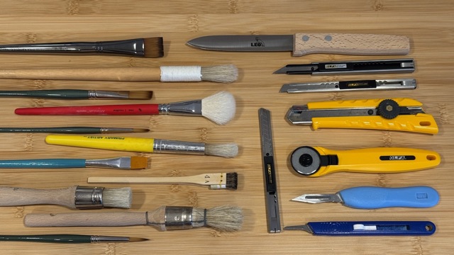 Paste and glue brushes of many sizes beside cutting tools on a bench.