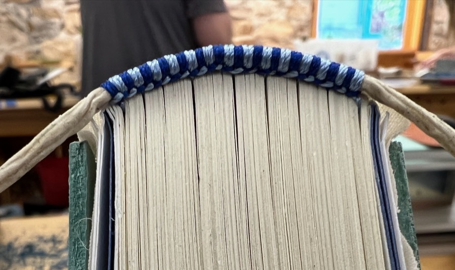 A silk endband being sewn onto the head of a text block held in a finishing press.